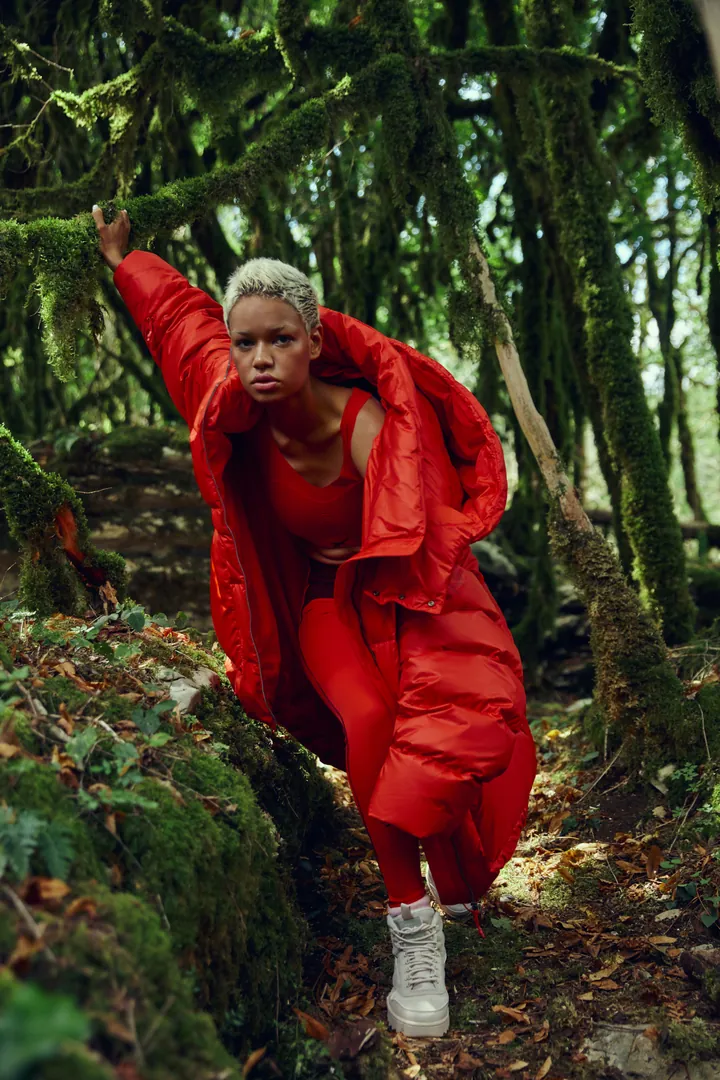 A model in a red down suit crouching in a mossy forest