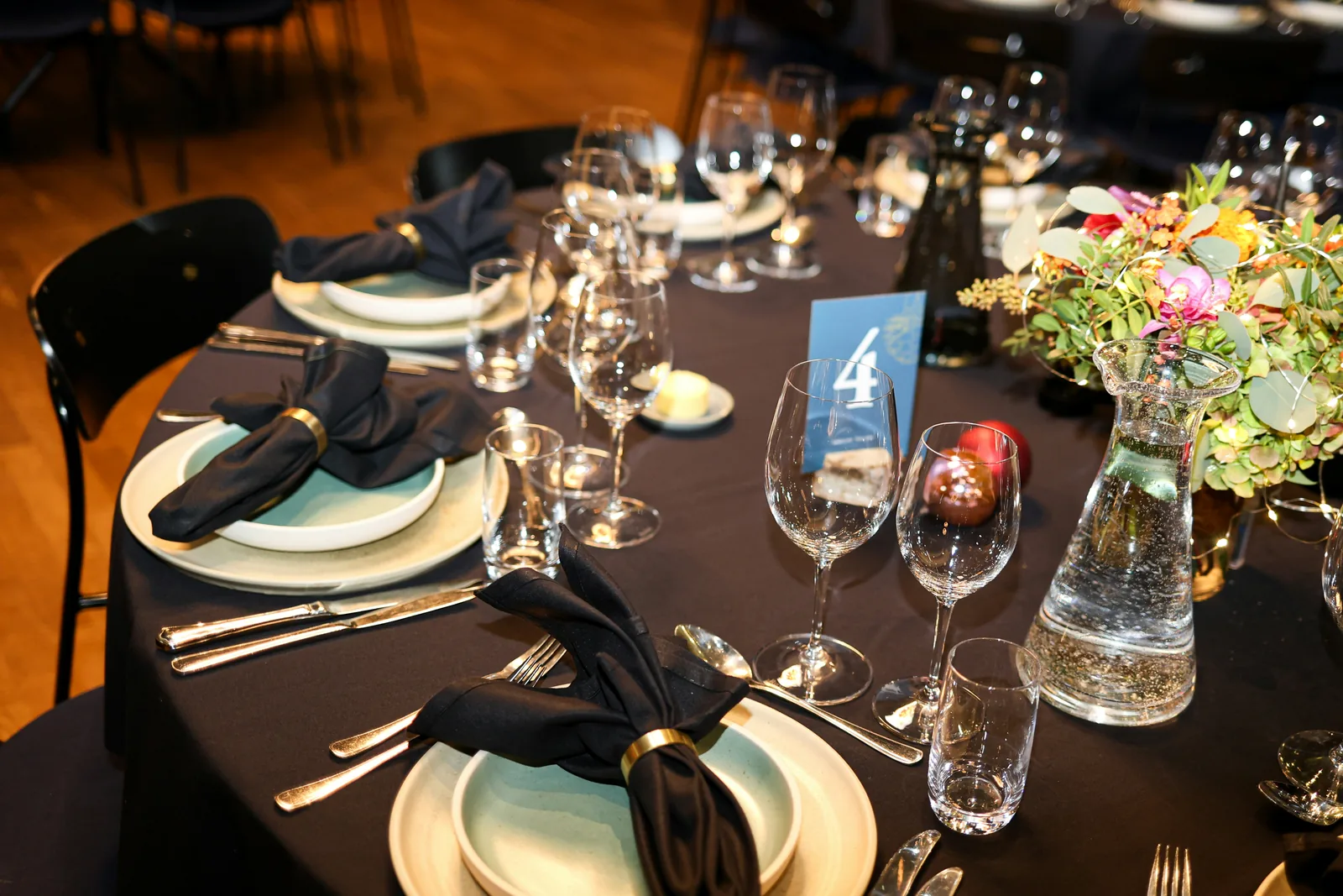 A round dinner table set for a corporate event: black linen, pale plates with black napkins in brass rings, crystal glasses, a water carafe, a blue card with table number 4 and a bright floral centrepiece, with more set tables behind.