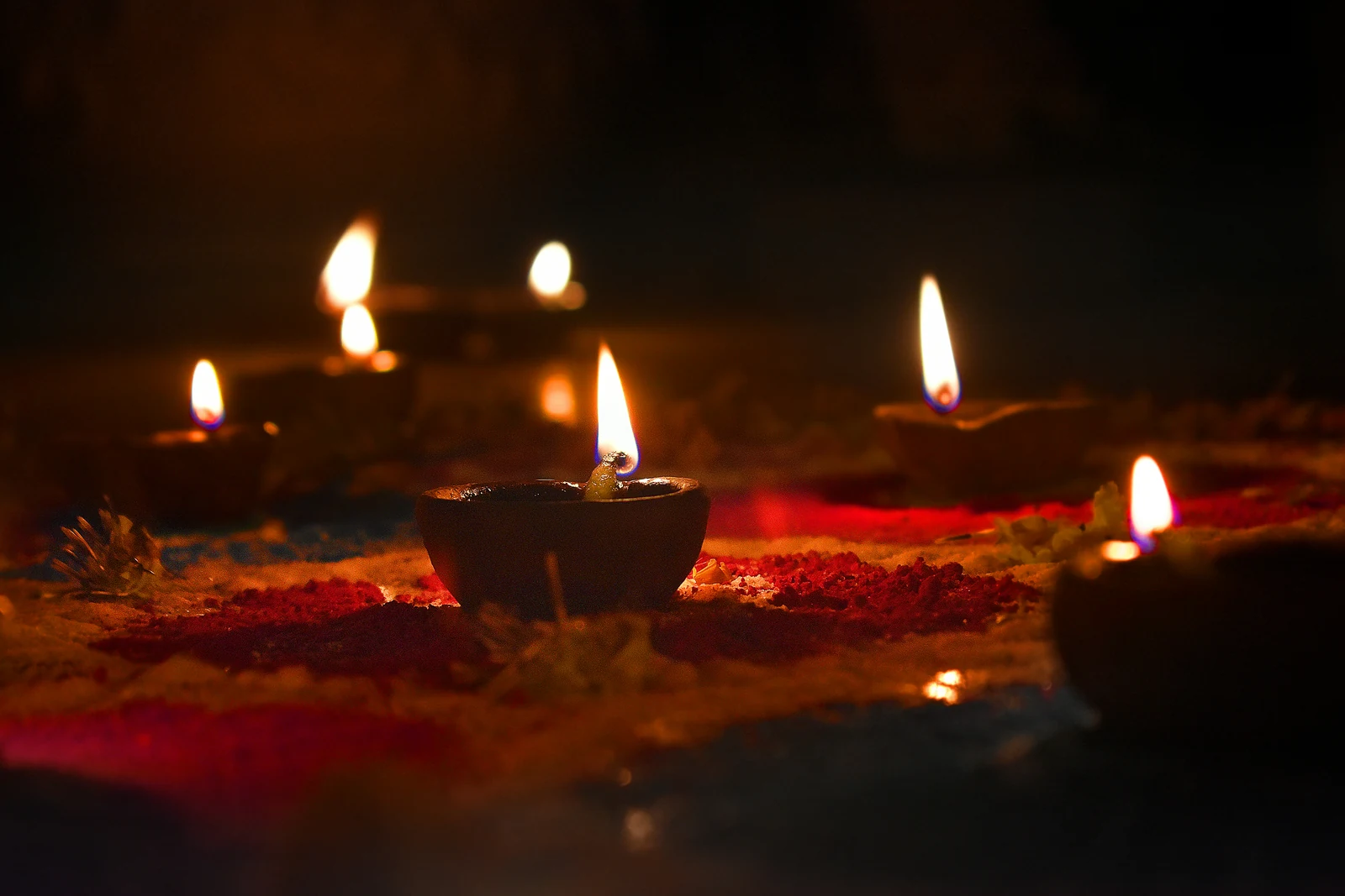 Clay diya oil lamps lit on a red and saffron rangoli in the dark, one flame in sharp focus and the rest blurred behind it.