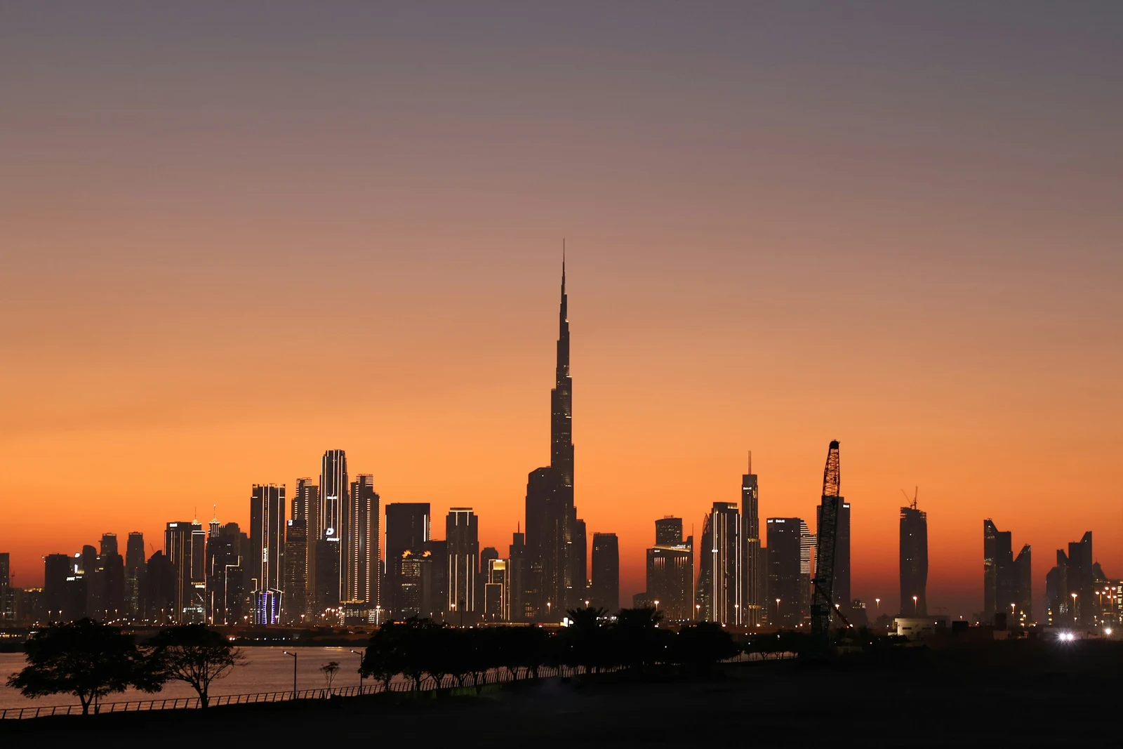 The Dubai skyline at sunset: Burj Khalifa rises above the downtown towers against an orange sky, with a row of trees and the waterfront in silhouette in front.