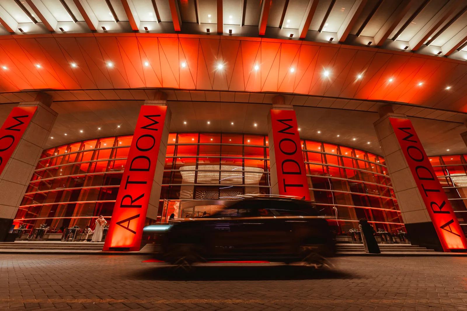 A car blurs past a venue entrance at night: red-lit columns wrapped in ARTDOM banners, a curved glass facade glowing orange behind them.